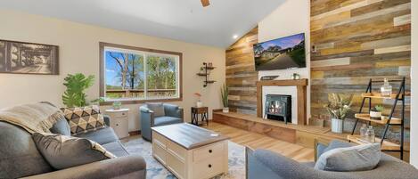 A modern living room with a fireplace, a ceiling fan, and a large window.