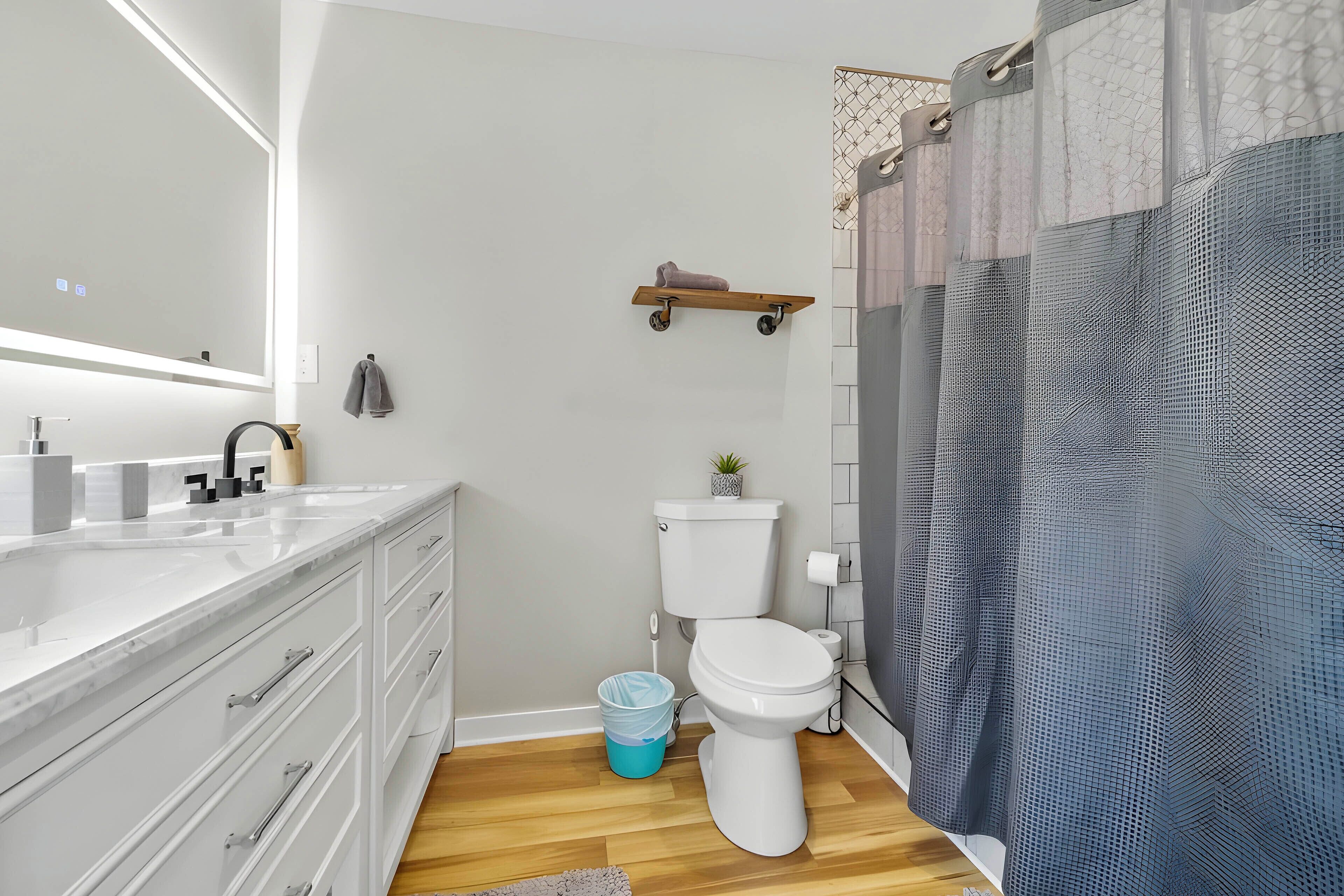 A modern bathroom with a white sink, mirror, and toilet. There is a wooden shelf above the toilet and a blue trash bin next to it.