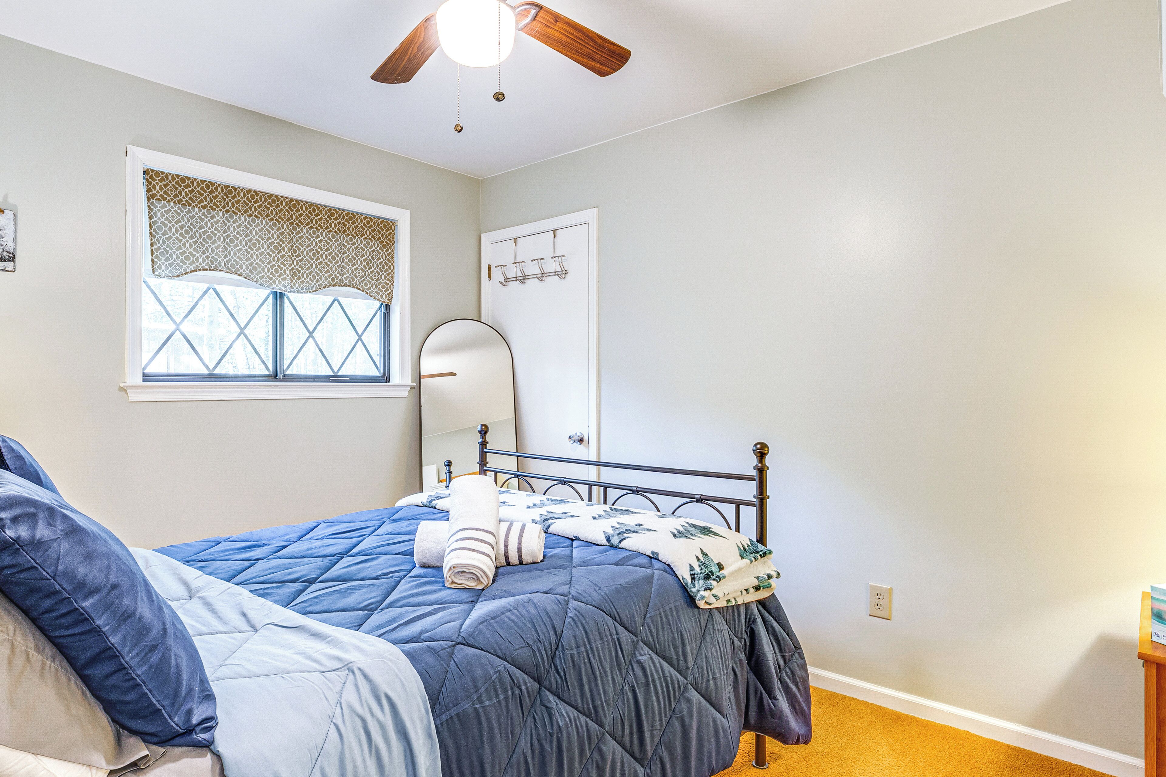 A bedroom with a bed, a ceiling fan, a window with a decorative valance, a door with a coat rack, and a small wooden nightstand.