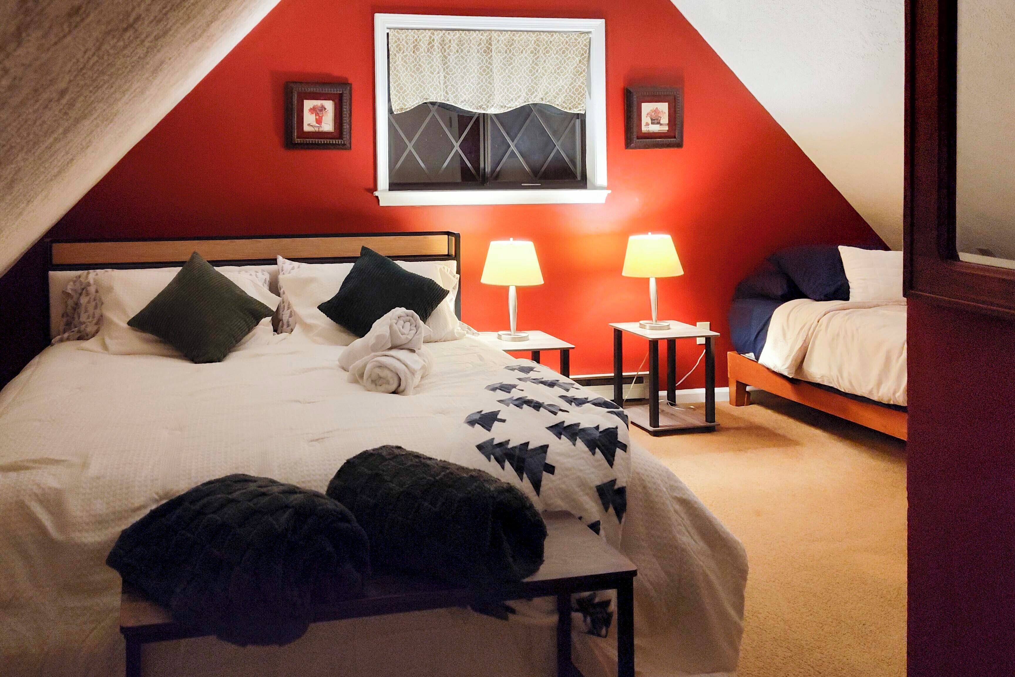 Een slaapkamer met een groot bed, twee nachtkastjes met lampen, een raam met een patroon gordijn, en een rode accentmuur.