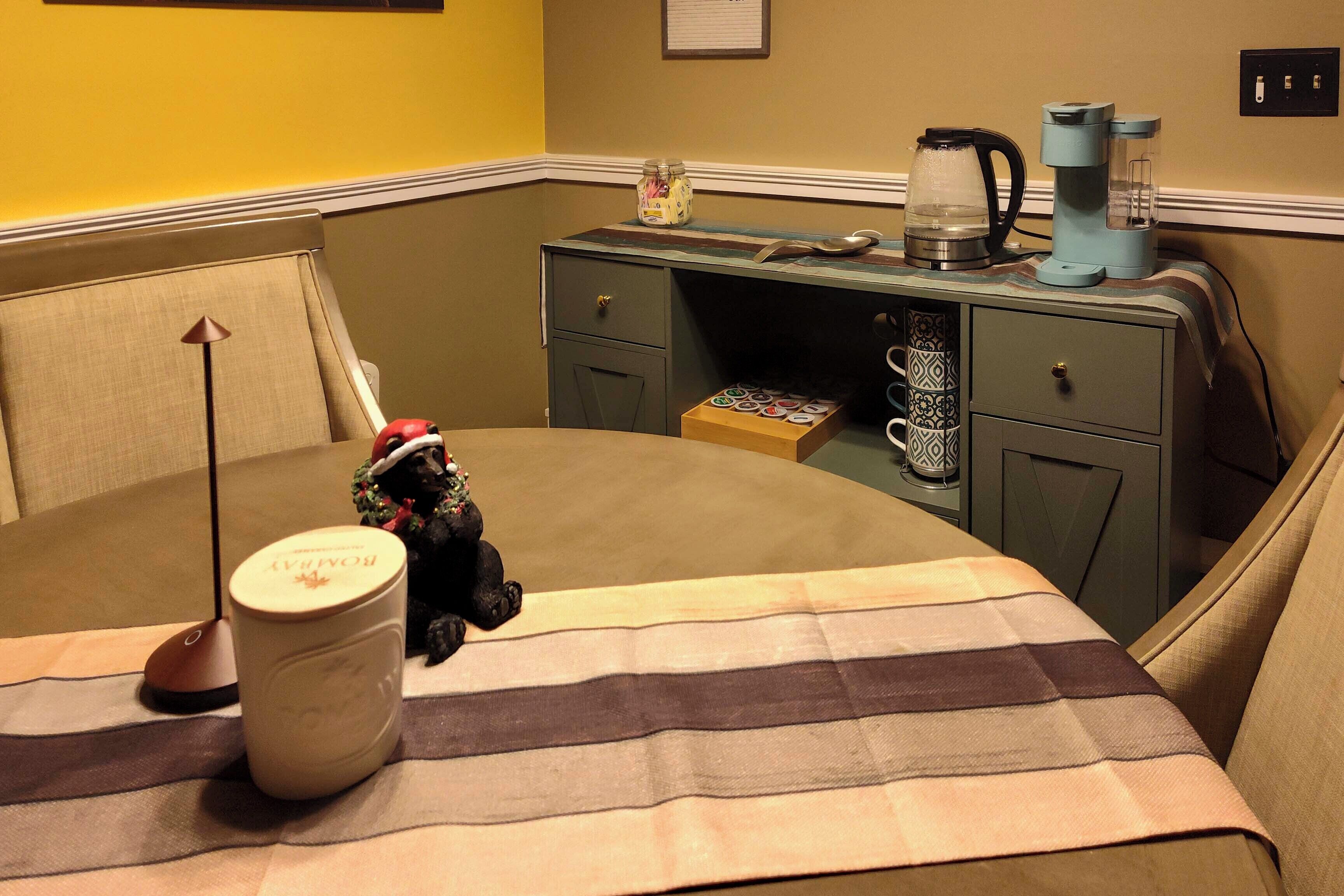 A cozy hotel room with a bed, a small desk with a coffee maker, a jar of snacks, and a decorative item.
