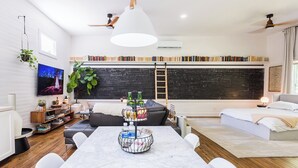 Una sala moderna con una amplia estantería, una mesa de comedor con portavasos para botellas de agua, un sofá gris y una cama con cabecera blanca.
