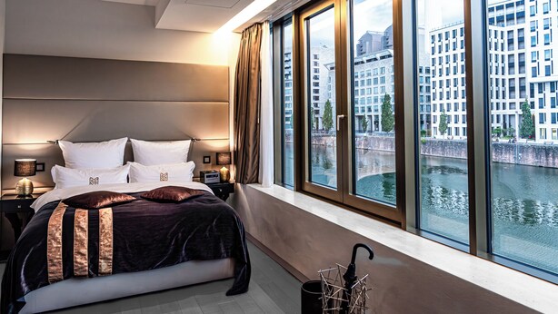 A modern hotel room with a large bed, a nightstand, a lamp, and a view of a river and buildings. - Main Suites (Frankfurt)