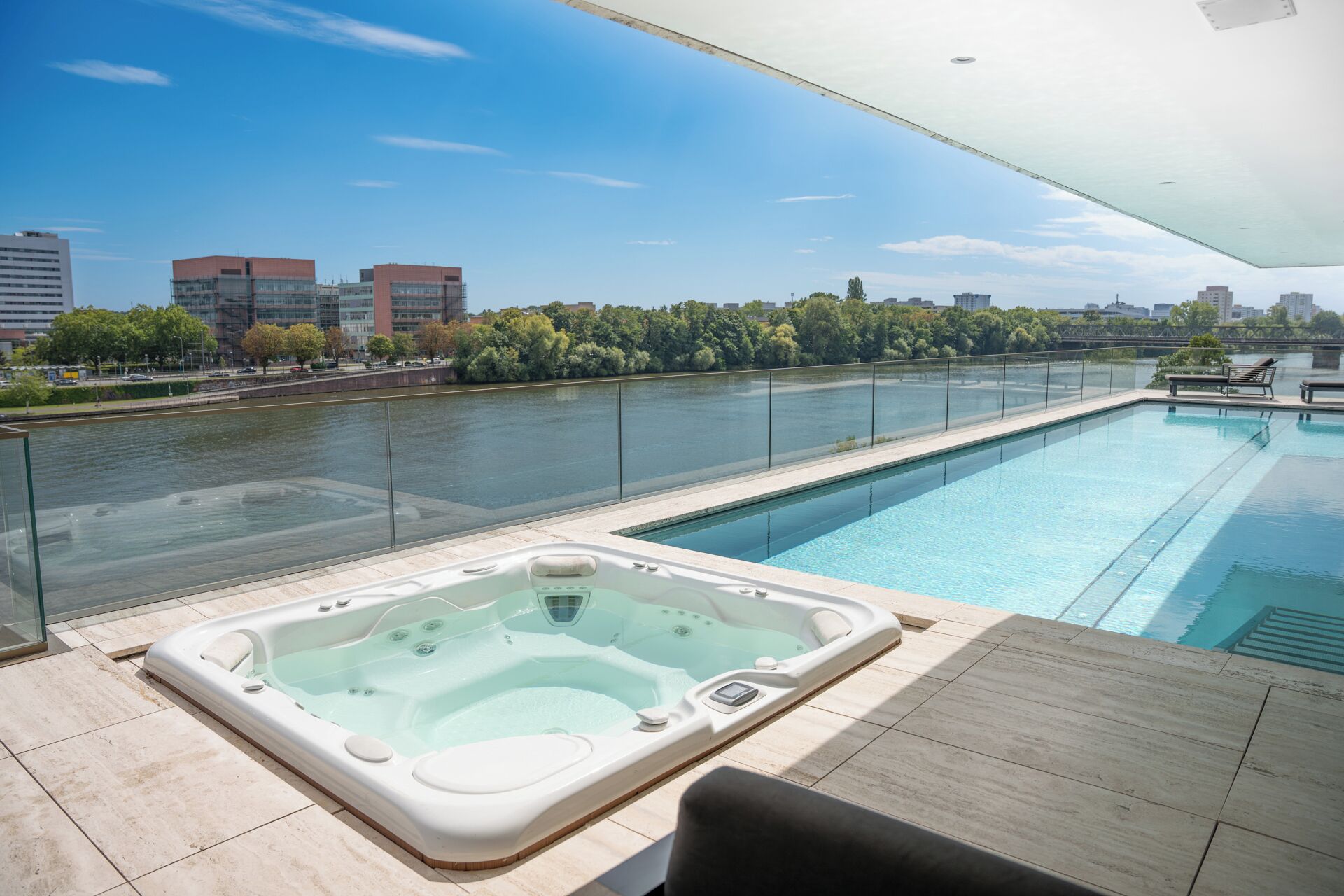 A rooftop swimming pool with a hot tub, surrounded by a glass railing, overlooking a river and cityscape.