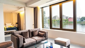 A modern hotel room with a sofa, a glass coffee table, and a view of a river and buildings. - Main Suites (Frankfurt)