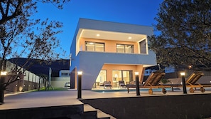 A modern two-story house with a swimming pool, outdoor seating, and a well-lit entrance.