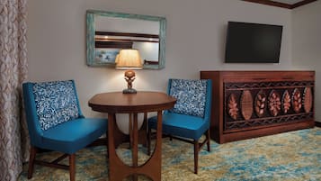 A room with two blue upholstered chairs, a small round table with a lamp, a mirror, and a flat-screen TV mounted on the wall.