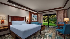 A hotel room with a large bed, a sofa, a desk with a chair, and a view of a garden. - Disney's Polynesian Villas & Bungalows (Lake Buena Vista)