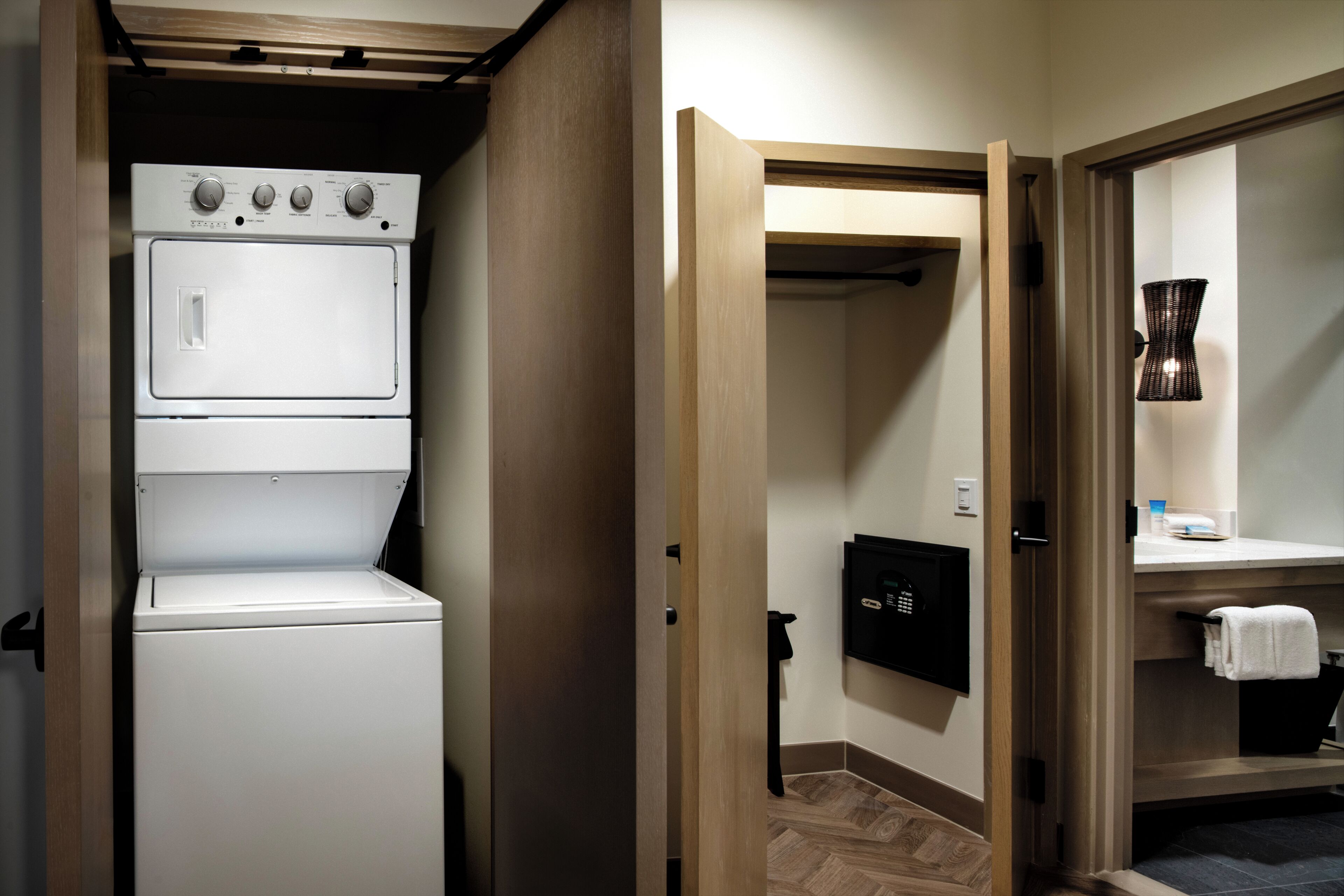 Dans une chambre d’hôtel, on trouve un espace buanderie avec une machine à laver et un sèche-linge superposés, un panneau de sécurité noir et un porte-serviettes.