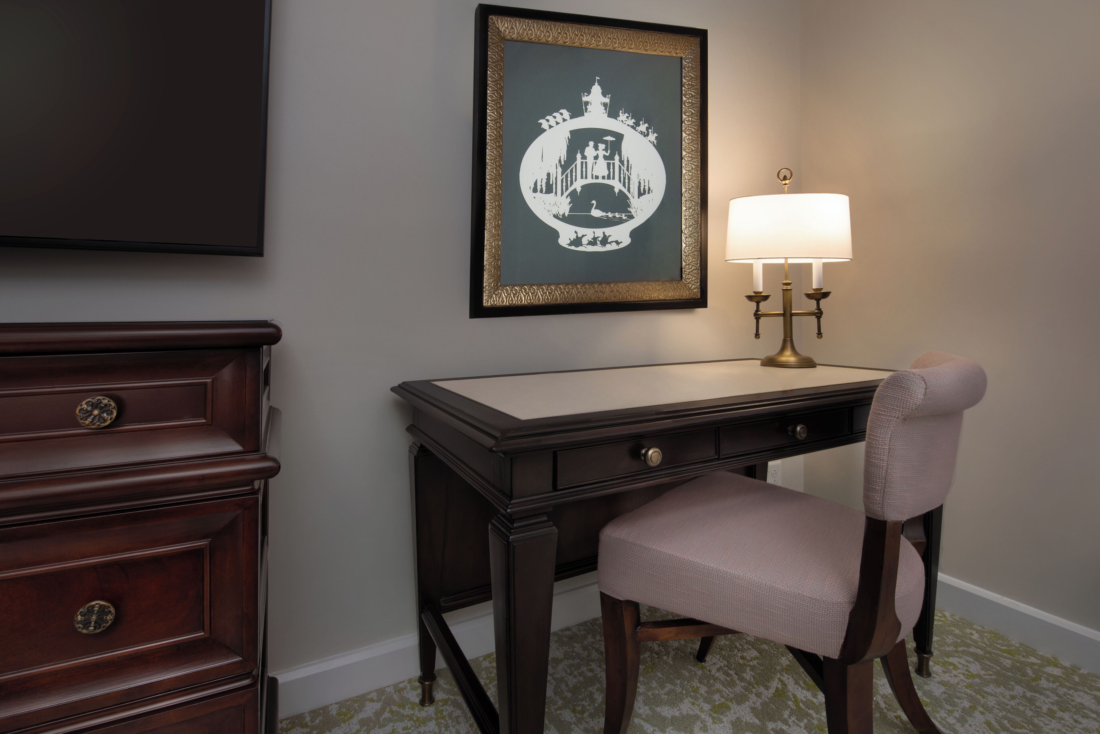 A dark wood desk with a lamp, a framed picture, and a chair.