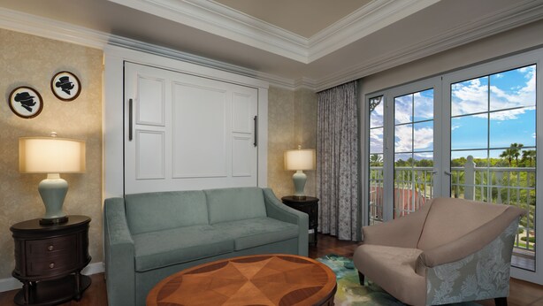 A room with a sofa, armchair, and a round table. There are two lamps, a door, and a window with a view. - The Villas At Disney's Grand Floridian Resort & Spa (Lake Buena Vista)