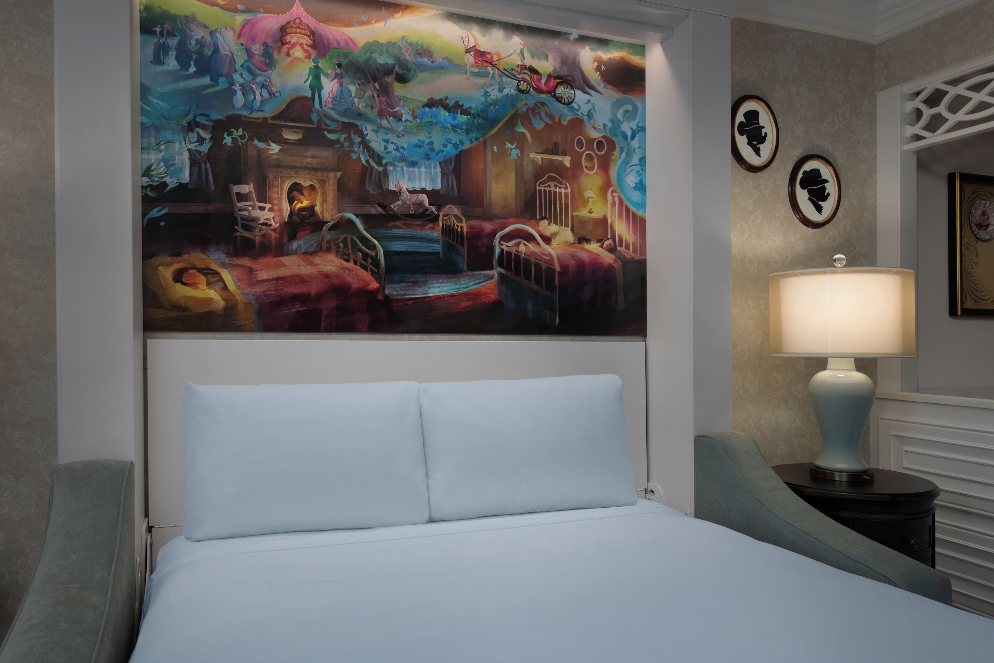 A hotel room with a large wall mural, a bed with white linens, a nightstand with a lamp, and a framed picture on the wall.