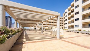 Un espacio al aire libre cubierto con suelo de baldosas, macetas de concreto con flores y un edificio de varios pisos al fondo.