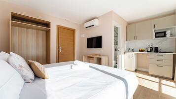A bedroom with a large bed, a wooden wardrobe, a TV, a small desk, and a kitchenette with a microwave.