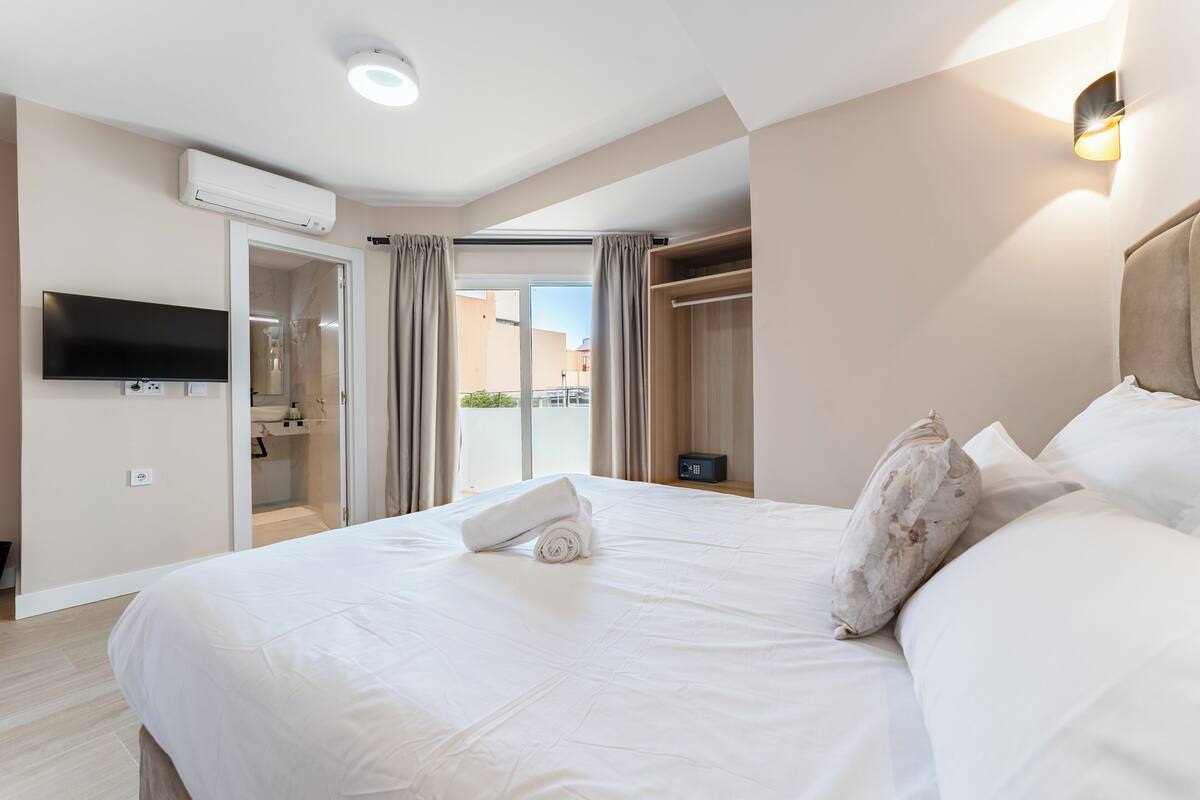 A hotel room with a large bed, a TV mounted on the wall, an air conditioning unit, a wardrobe, and a view of the outside through a glass door.