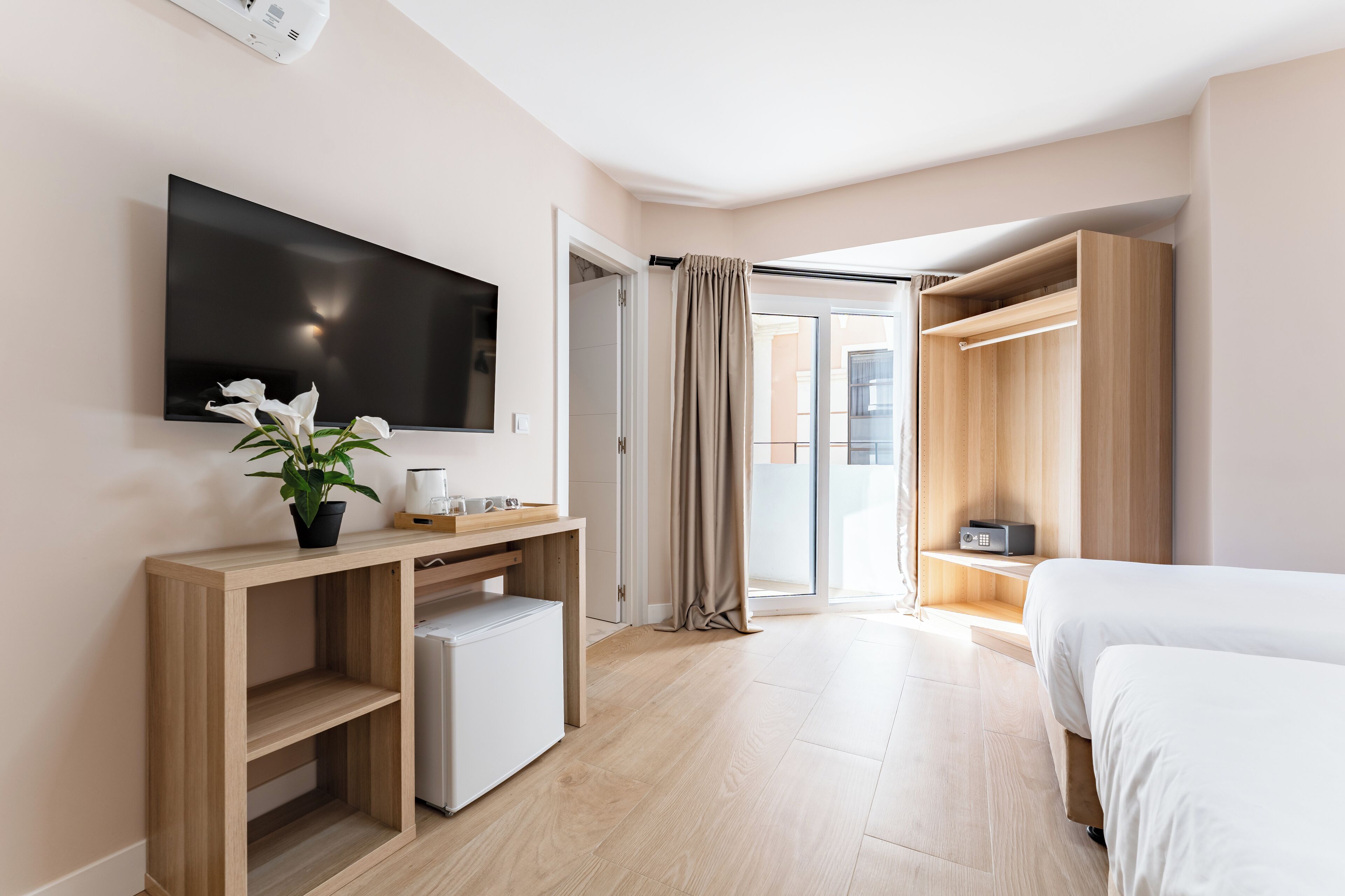 A modern hotel room with a flat-screen TV, a small desk with a plant, a mini-fridge, a bed, and a wardrobe.