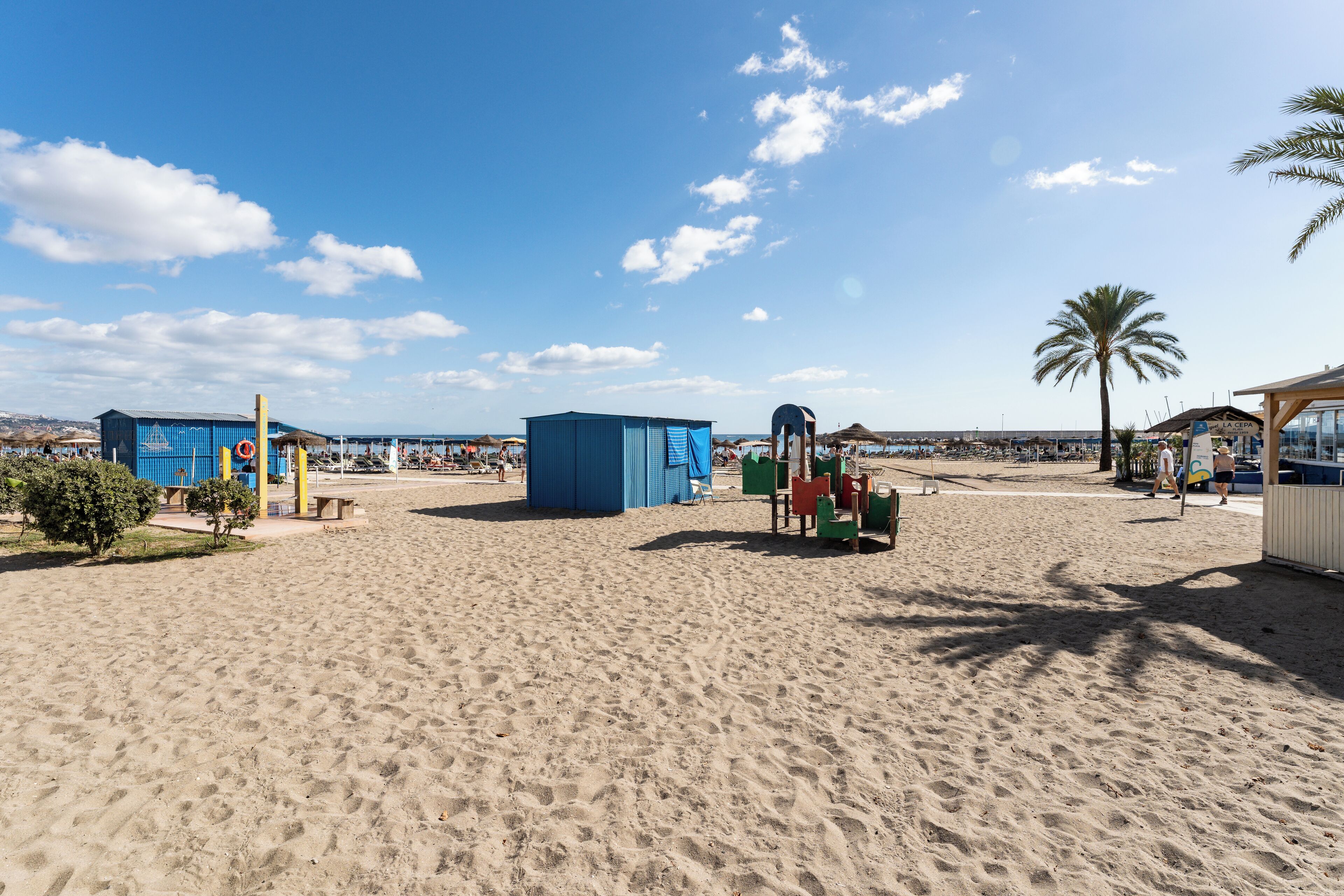 A sandy beach with blue and green structures, a palm tree, and a clear sky.