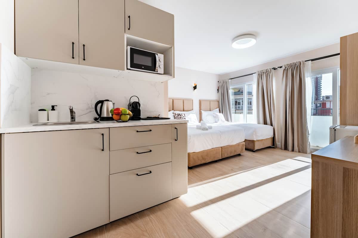A modern hotel room with a kitchenette, a bed with a headboard, a large window with curtains, and a view of the city.