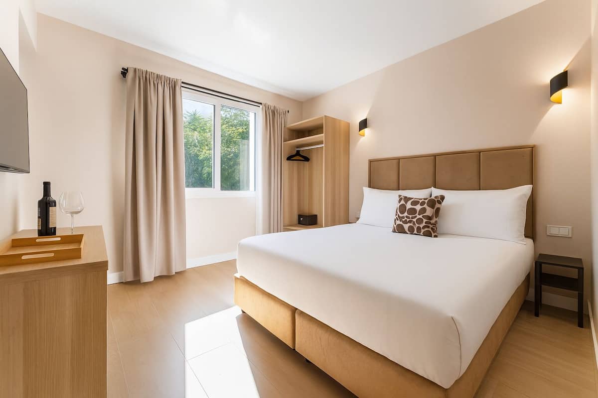 A modern hotel room with a large bed, a wardrobe, a bedside table, and a window with curtains.