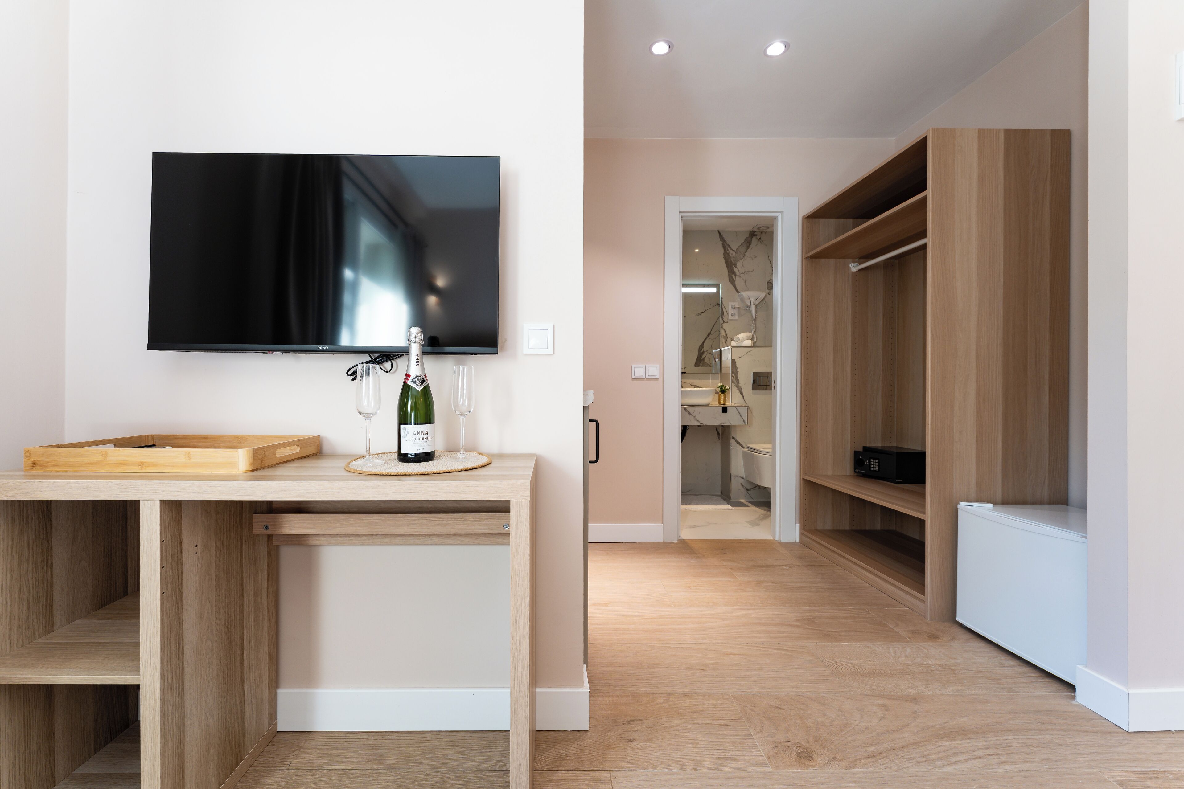 Un salon moderne avec un téléviseur à écran plat fixé au mur, une table basse en bois sur laquelle se trouvent une bouteille de vin et un verre, et un meuble encastré en bois doté d’étagères.