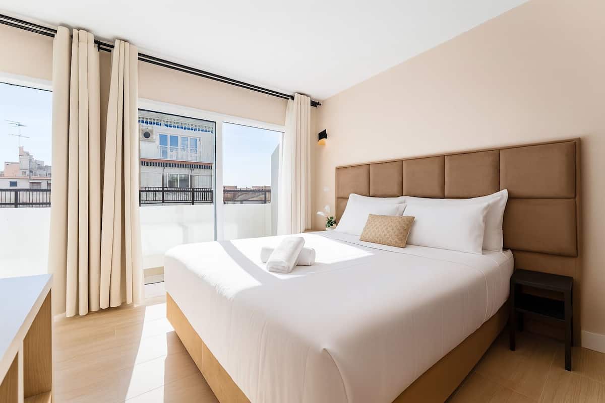 A hotel room with a large bed, a small bedside table, a view of the city through a balcony, and a window with curtains.