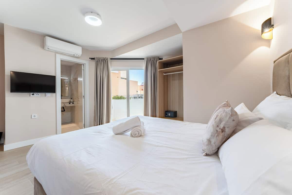 A hotel room with a large bed, a TV mounted on the wall, an air conditioning unit, a wardrobe, and a view of the outside through a glass door.