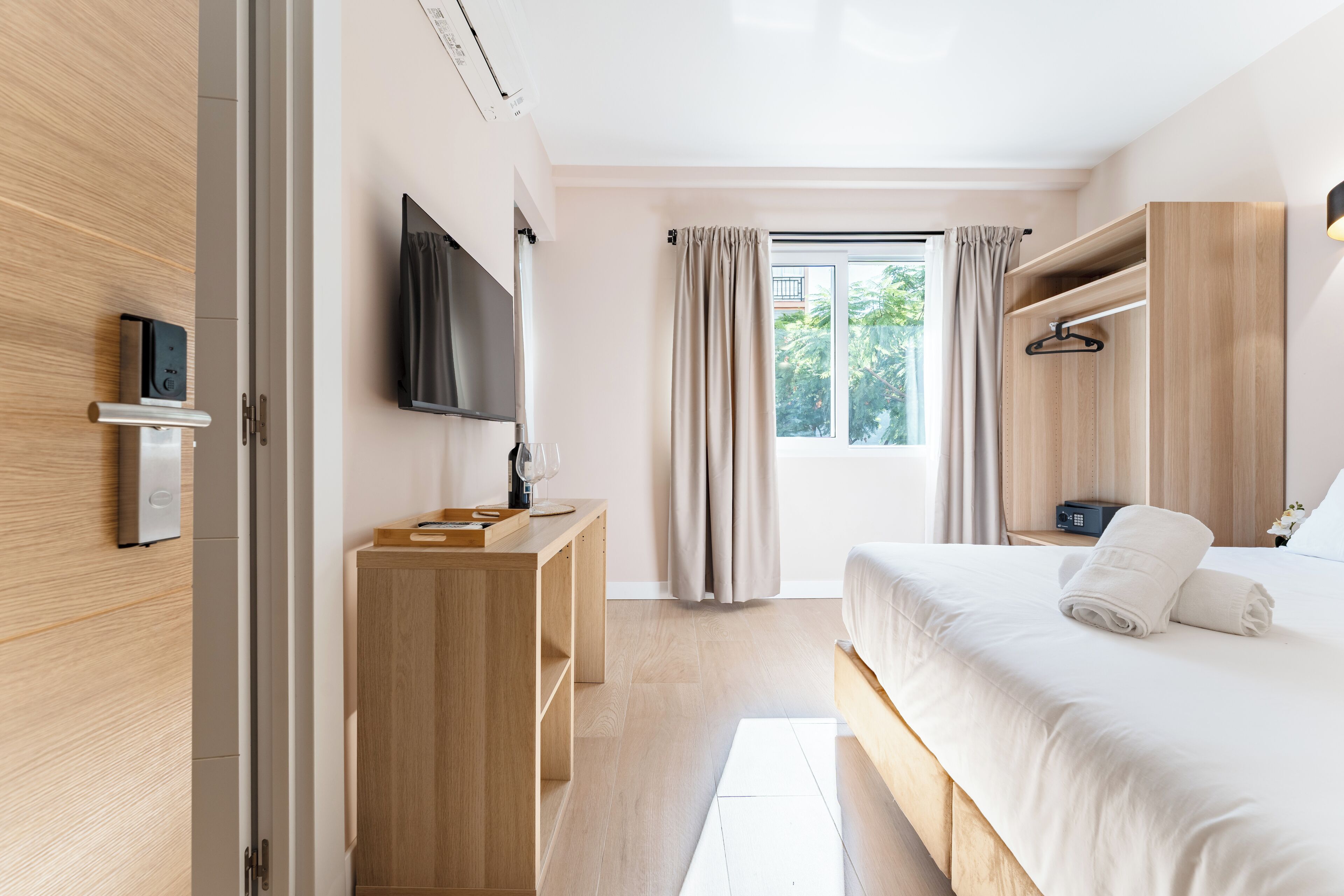 Habitación de hotel moderna con armario de madera, televisión de pantalla plana, mesita de noche, cama con ropa de cama blanca y ventana con cortinas.