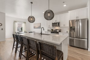 A modern kitchen with white cabinets, a stainless steel refrigerator, a large island with bar stools, and pendant lighting.