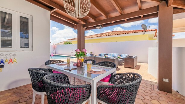 Un patio al aire libre con una mesa lista para comer, sillas de mimbre, una lámpara colgante y una vista de una piscina y un edificio al fondo.
