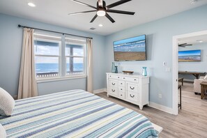 Ein Schlafzimmer mit einem Bett, einem Deckenventilator, einem Schminktisch und einem großen Fenster mit Blick auf den Ozean.