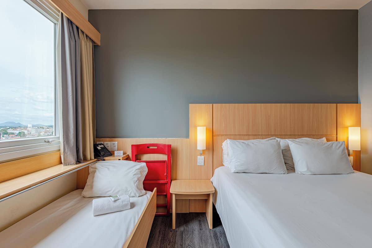 Ein Hotelzimmer mit zwei Betten, einem kleinen Tisch, einem roten Sessel und einem Fenster mit Vorhängen.