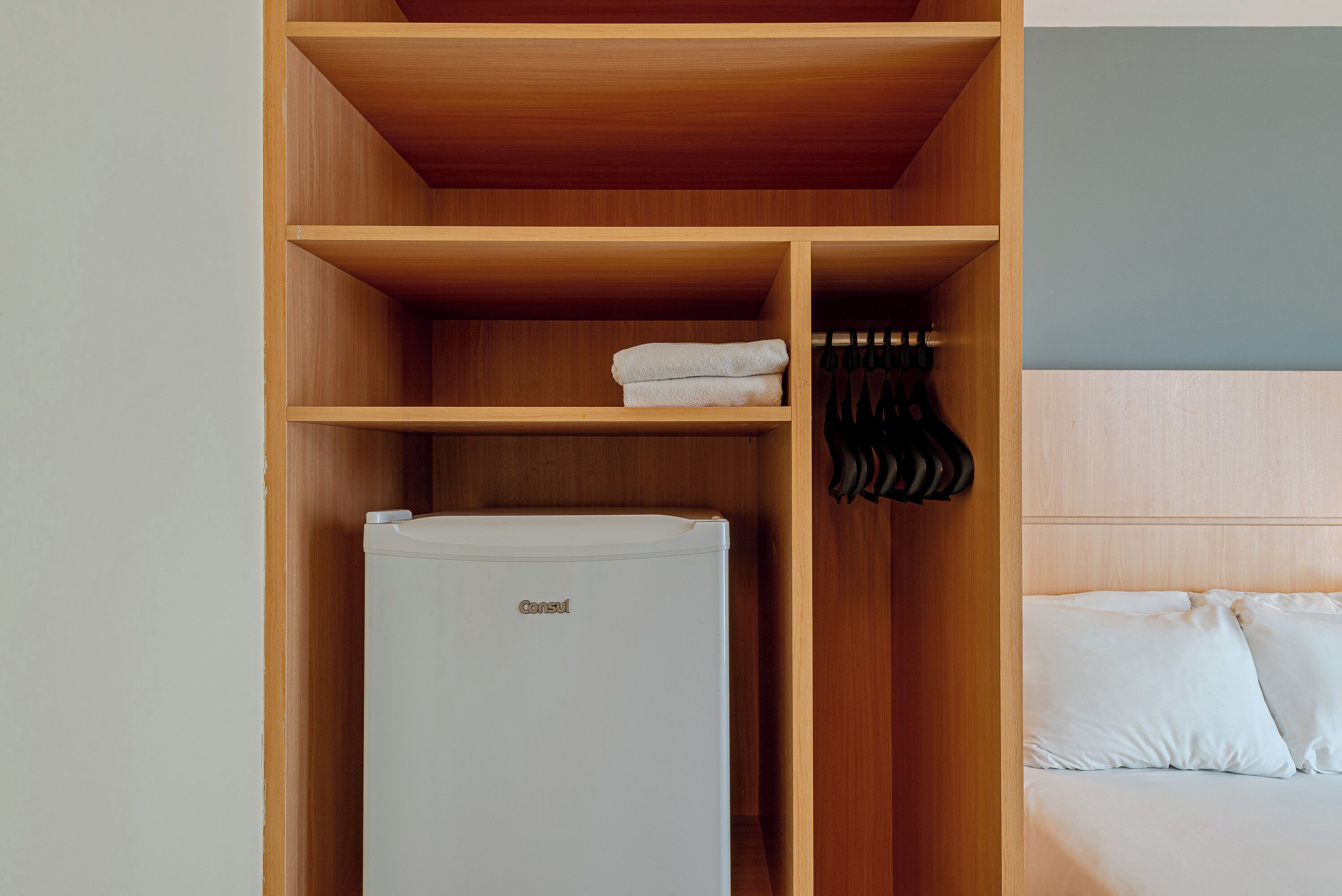 Una habitación de hotel con un armario de madera, una cama blanca y un refrigerador blanco.