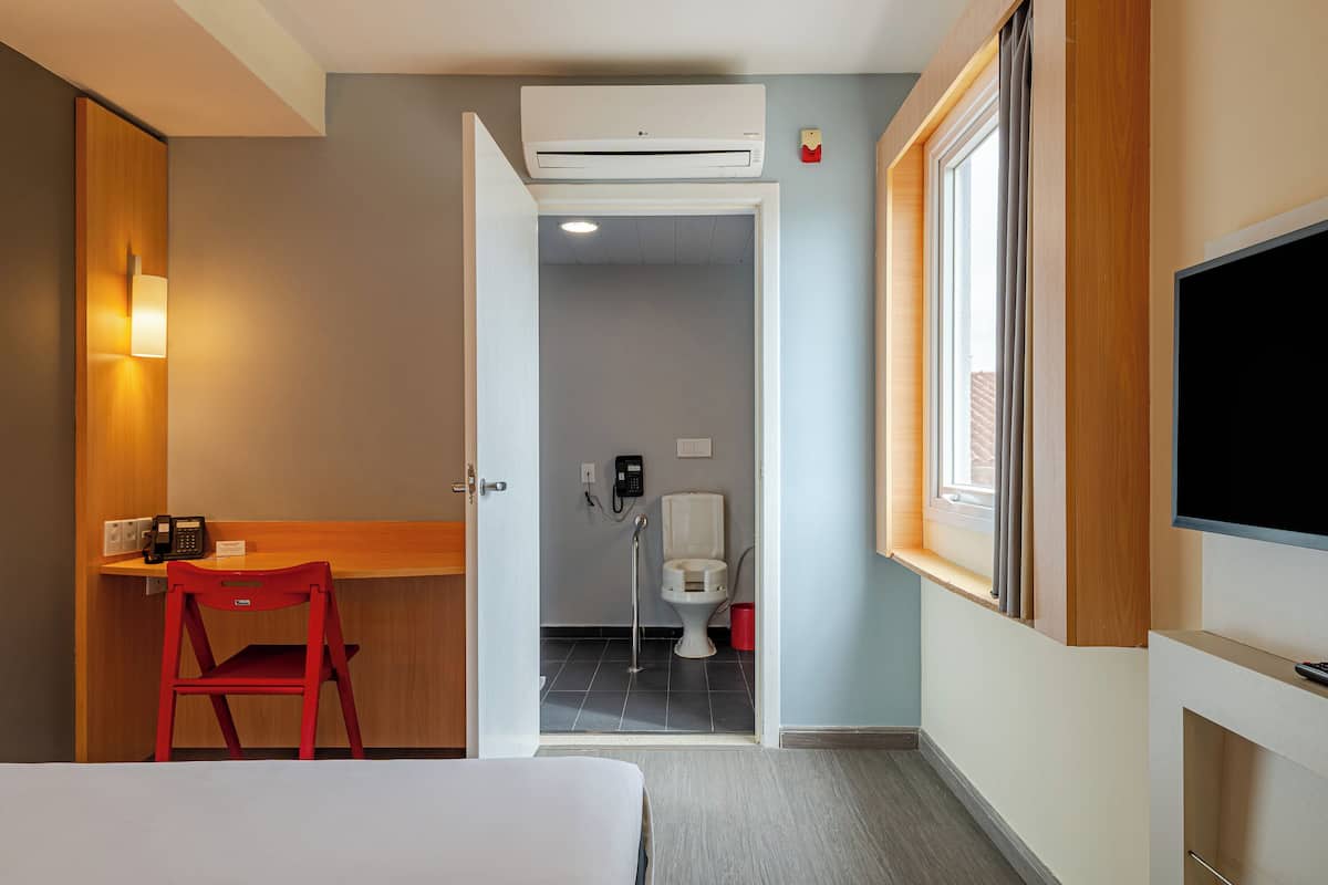 Ein Hotelzimmer mit einem Bett, einem Schreibtisch mit Stuhl, einem Fernseher, einer Klimaanlage und einem sichtbaren Badezimmer durch eine offene Tür.