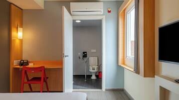 Habitación de hotel con una cama, un escritorio con silla, televisión, aire acondicionado y un baño visible a través de una puerta abierta.