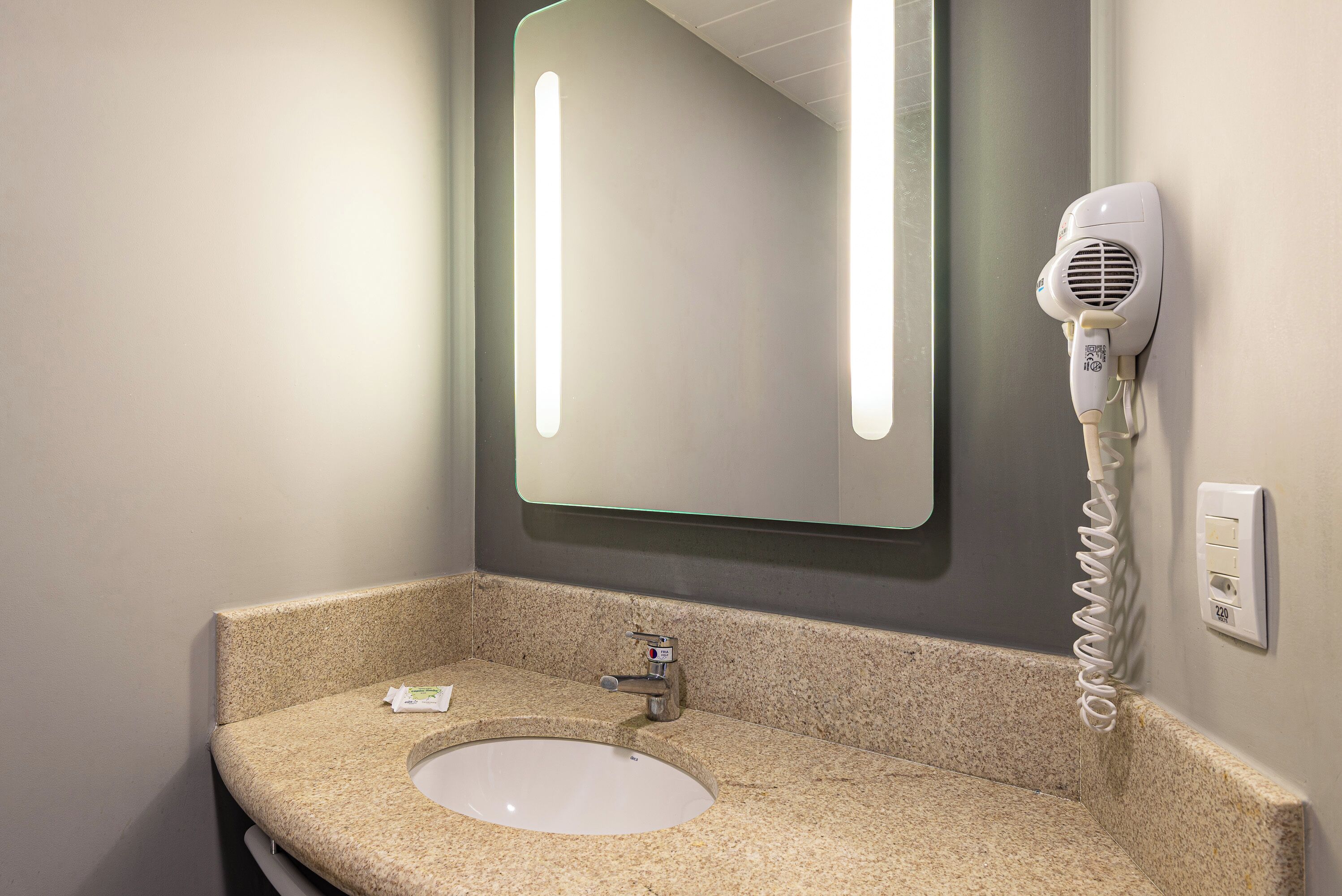 Un baño con encimera de granito, un lavabo, un espejo y un secador de pelo montado en la pared.
