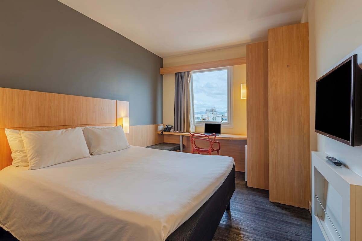 Ein Hotelzimmer mit einem großen Bett, einem Schreibtisch mit Computer, einem Fernseher und einem Fenster mit Vorhängen.