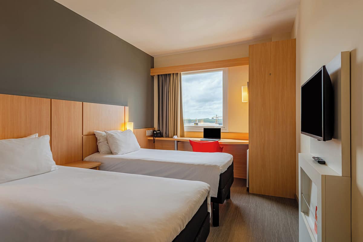 Ein Hotelzimmer mit zwei Betten, einem Schreibtisch mit Laptop, einem Fernseher und einem Fenster mit Blick.