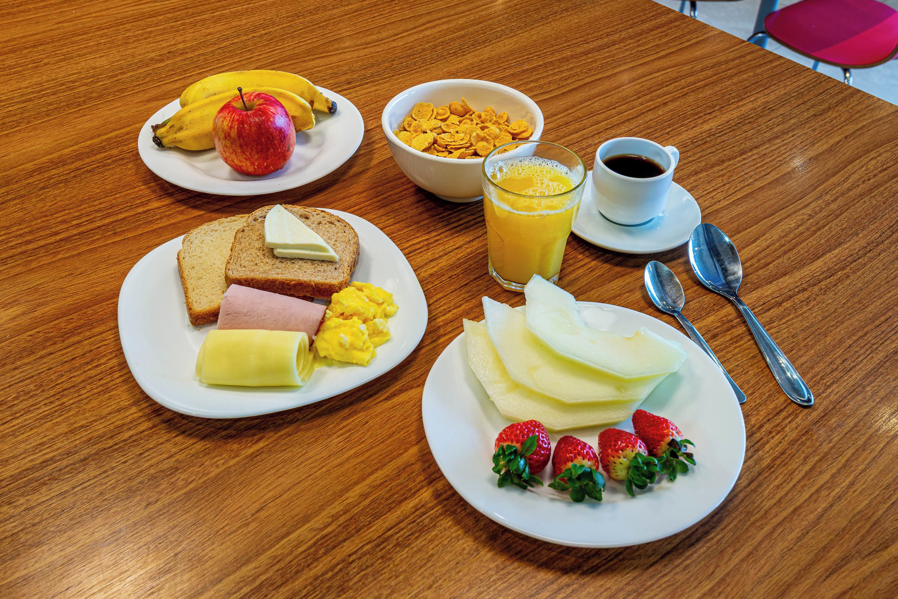 Et træbord med tallerkener med morgenmad, bestående af ristet brød, æg, skinke, ost og jordbær. Der er også bananer, et æble, en skål med cornflakes og et glas appelsinjuice samt en kop kaffe.