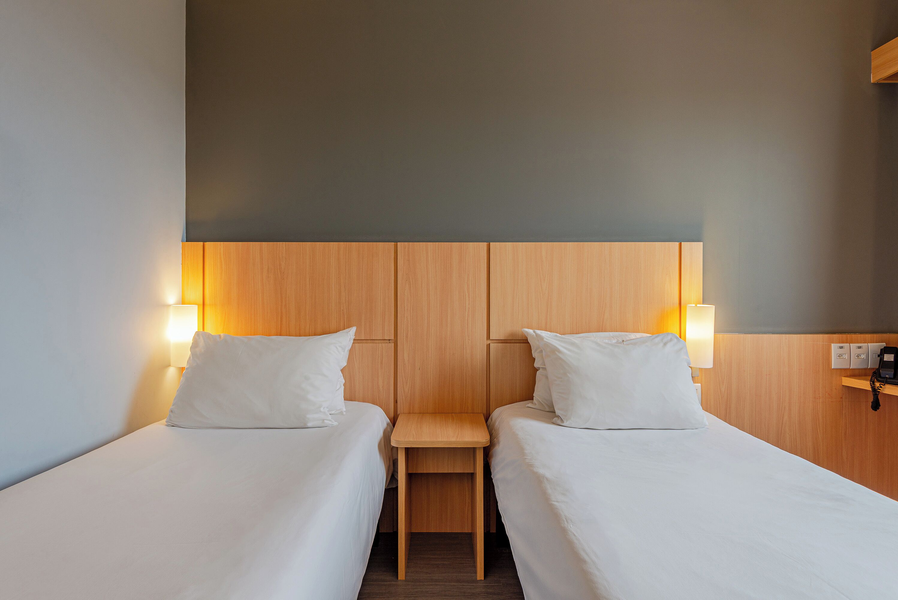 Una habitación de hotel con dos camas individuales, una mesita de noche de madera y una lámpara fijada a la pared.