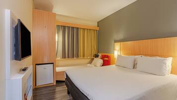 Quarto de hotel com cama, televisão, mini-geladeira e escrivaninha com luminária.