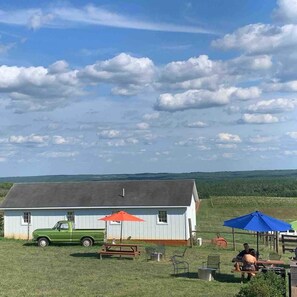 Un cadre extérieur rustique avec un bâtiment blanc, un camion vert, des tables de pique-nique et un parasol bleu.