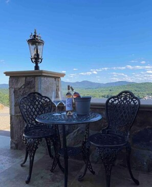 Un espace extérieur aménagé avec deux chaises en métal décorées et une petite table ronde, une bouteille de vin et un verre. On aperçoit une lanterne en pierre à l’arrière-plan.