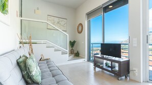 Un salon moderne avec un canapé gris, un téléviseur posé sur un meuble, un escalier et une grande fenêtre donnant sur un beau paysage.