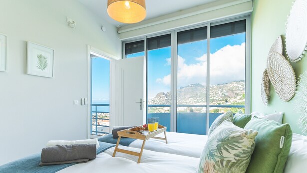 Une chambre avec une grande fenêtre, un lit sur lequel est posé un plateau de petit-déjeuner, et une vue sur un paysage côtier.