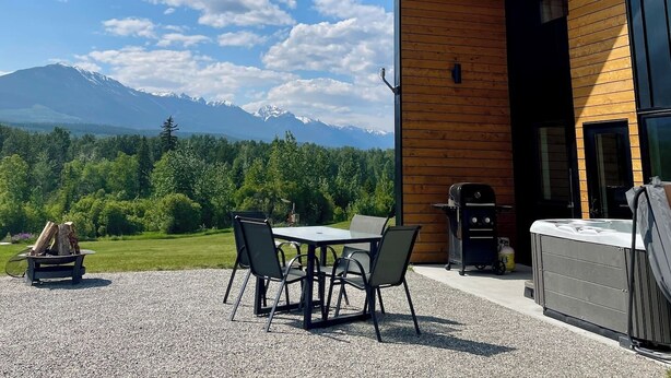 A modern building with a sloped roof and large windows, surrounded by a gravel patio with a table and chairs, and a view of a mountain range. - Epic Mountain View | Private Hot Tub | PÜR 1 (Golden)