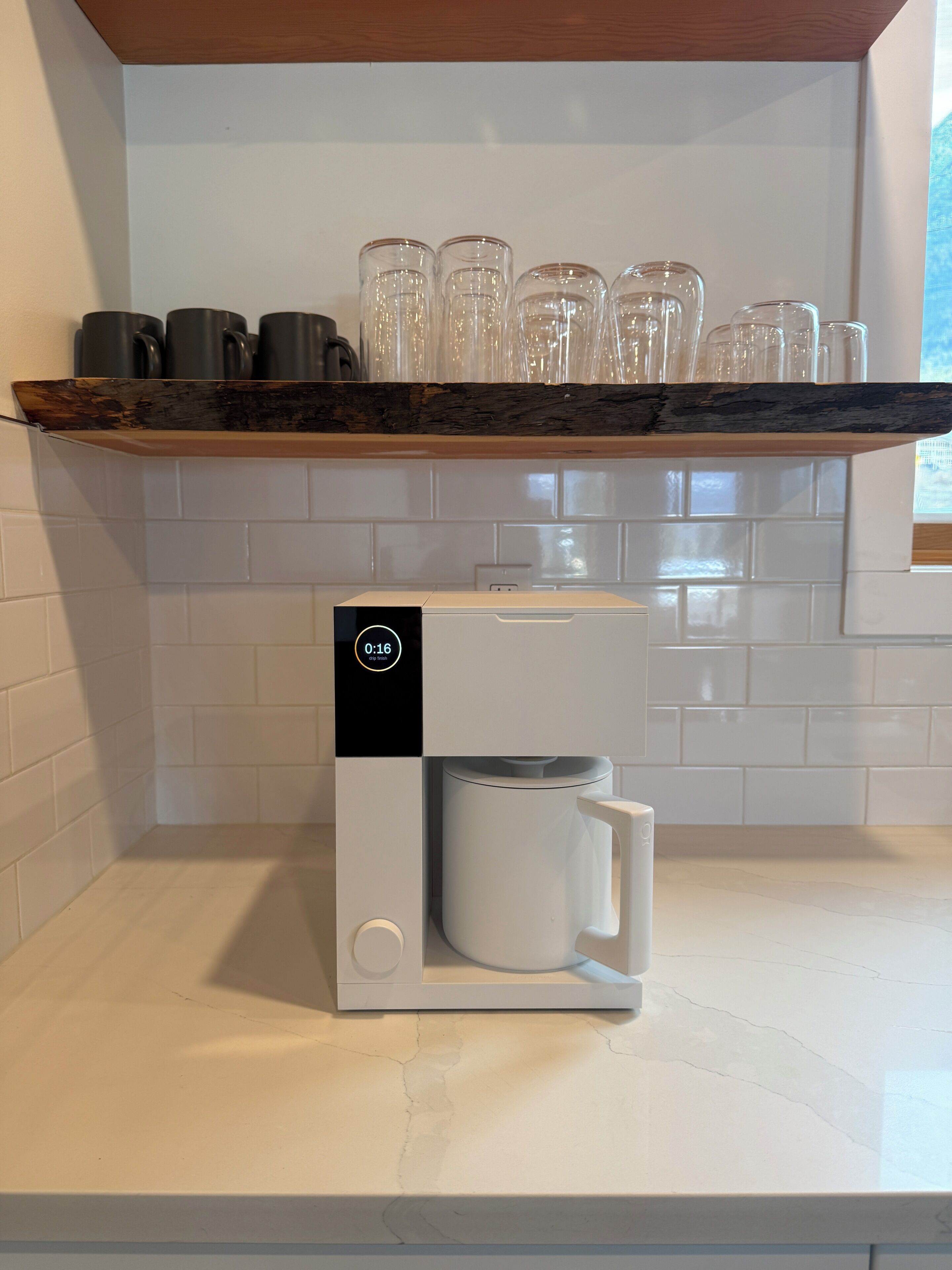 A kitchen with a coffee machine, a shelf with glasses, and a window.
