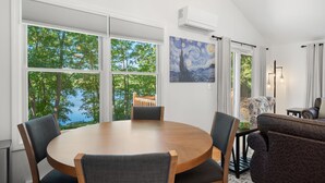 Un comedor con una mesa redonda y sillas, ventanas amplias con vistas a árboles y una masa de agua, un cuadro en la pared y un ventilador de techo.