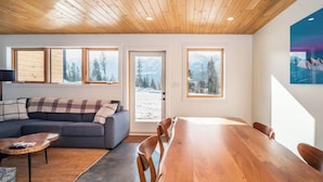 A modern living room with a wooden ceiling, a grey sofa with plaid cushions, a wooden dining table, and large windows with snowy mountain views. - Unreal Mountain Views | Private Hot Tub | PÜR 3 (Golden)