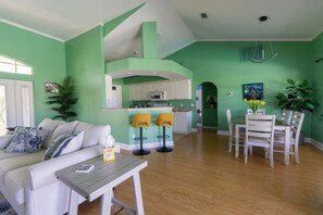 A living room with a sofa, a coffee table, and a dining area with a table and chairs. There is a kitchen area with white cabinets and a bar counter. - Island Time (Cape Coral)