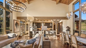 Un amplio salón con un sofá grande, una mesa de comedor y sillas, vigas de madera y ventanas grandes que ofrecen vistas al exterior.